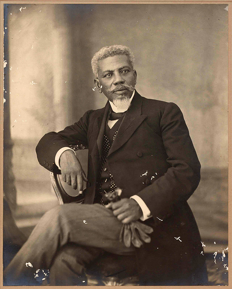 A black man with handlebar mustache and goatee Faces te camer but is seated in three-quarter profile, left leg crossed over right. He is wearing a three-piece suit, bowtie and holds a pair of gloves in his left hand, resting on his leg. The caption in the source reads "Cincinnatus Leconte, President of Haiti. 1911-1912. Haitian Collection,1775-1950. Library of Congress Manuscript Division."