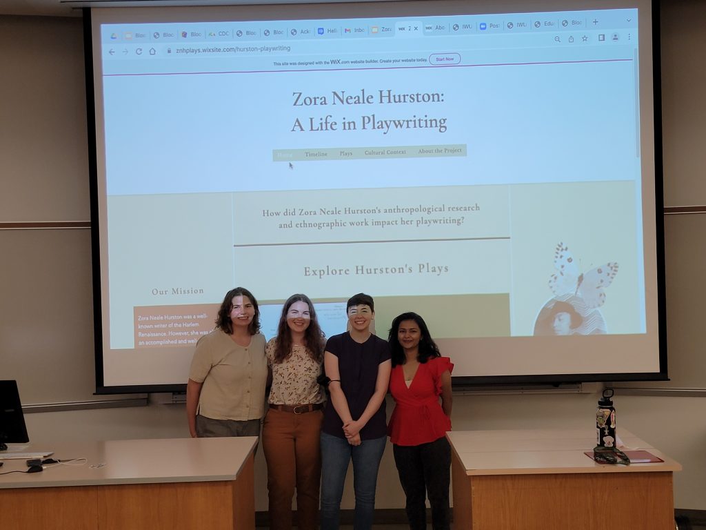 Digital Humanities Fellows Mishwa Bhavsar, Leah Rosen, Julia McMahon and Ellie Kurtz stand in front of a screen with their project's website projected on it.