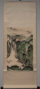 Black ink wash Chinese scroll painting; color landscape; cloud-shrouded mountains span the top; a long series of mountain passes, some colored green, are divided by a river that descends from upper left into a long waterfall in middle-to-lower left third of this work; several figures are in the foreground on a rise of land by the water; several red-roofed buildings are visible in the upper right; a pathway with some stairs are visible in the lower right.